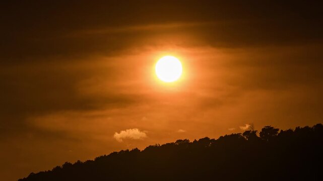 Time-lapse of the sun disappearing behind dark mountain silhouettes under a majestic sky.