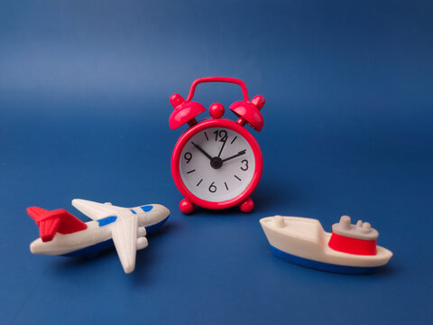 red vintage alarm clock stands between a small toy airplane and a miniature cargo ship on blue background