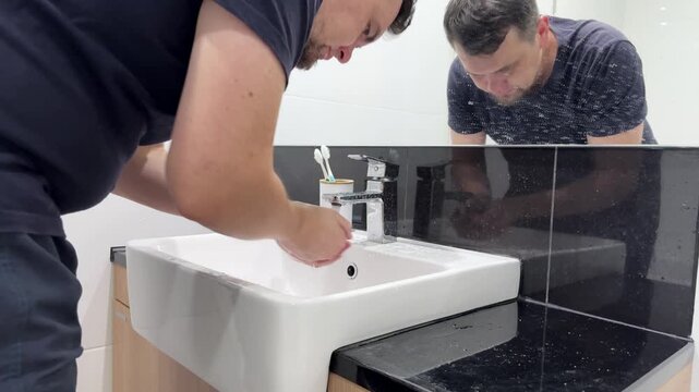 A man washes his face in the bathroom sink while preparing for the day.