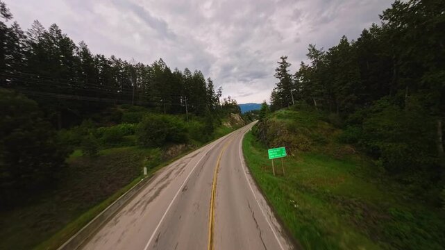 First-person view drone flies low and fast along a curving two-lane rural asphalt highway past a green distance sign before gradually ascending to reveal a straight stretch of road cutting through den
