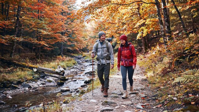 Happy couple with backpacks and trekking poles walking on path in beautiful autumn forest with colorful leaves near river.