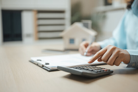 Person using a calculator to estimate mortgage costs with documents and a house model in the background. Concept of home loan planning, interest rate calculation, housing affordability.