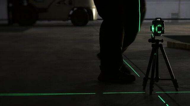 Technician kneels to adjust a green laser level on an industrial warehouse floor before construction, installation or alignment work. Precision measurement equipment in use.
