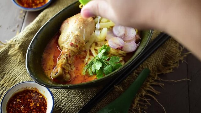 Hand squeezing fresh lime juice over Khao Soi, popular Northern Thai chicken curry noodle soup, adding sour flavor to delicious spicy Asian food culture dish.