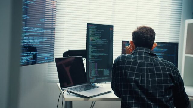 Young Caucasian male programmer writes code using multiple screens in home office workspace. AI development, cybersecurity, network database system, software engineering, remote tech startup concept.