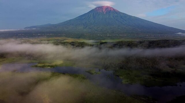 Aerial view of Mount Kerinci volcano rising above morning mist and lush green fields in Kayu Aro, Jambi, Indonesia.