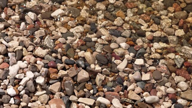 Close-up of crystal clear water flowing smoothly over a bed of small, colorful pebbles and stones, creating gentle ripples and reflections on the surface for a calming natural background