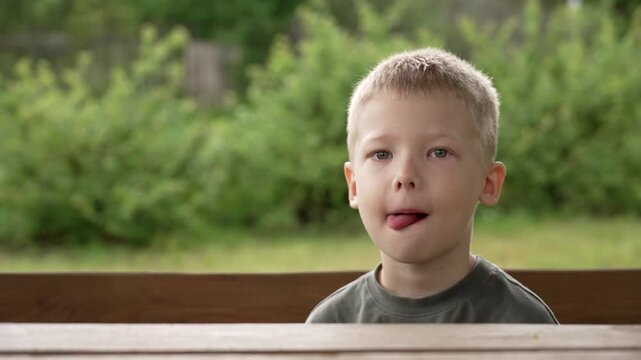 Adorable little boy with blond hair sitting at a wooden table outdoors, playfully sticking out his tongue and making a series of funny, silly faces while looking directly at the camera