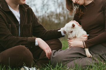 Obraz premium A couple sits on grass, gently holding a small goat kid between them, sharing a quiet moment outdoors, with soft natural light and a warm, intimate mood focused on care and companionship.