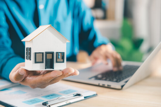 Real estate contract property mortgage finance approved insurance home document for homeowner tenant investment shows hand holding house model while using laptop computer at office desk