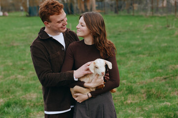 Obraz premium Couple, woman, man, goat, kid, outdoors, lifestyle, affection, animals, romantic. A smiling pair in a grassy park cradle a gentle goat kid, capturing a warm candid moment of tenderness and connection