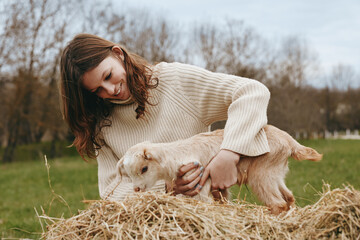 Obraz premium Joyful woman outdoors, gently holding a goat kid on hay, warm natural light, cozy sweater, rustic farm setting, tender moment of connection and care, nature vibes