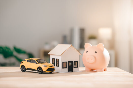 Financial wealth, savings, and growth shown by pink piggy bank, model house, and yellow car on wooden table represent real estate asset construction or debt management plans for future