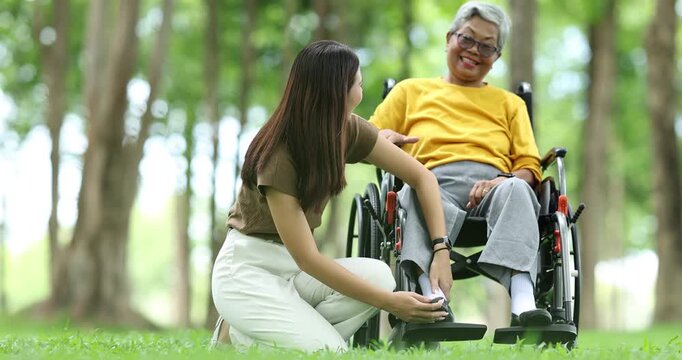 Young caregiver providing comforting support to an elderly asian woman using a wheelchair while enjoying a peaceful day outdoors in a green park, focusing on assistance and senior care