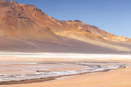 View of the vast salt flats of Salar de Aguas Calientes with arid, colorful volcanic mountains under a clear blue sky in San Pedro de Atacama, Antofagasta, Chile.