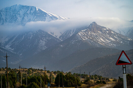 View of the snow-capped peaks of the Zaamin Mountains with low-hanging clouds, utility poles, and a road sign in the foreground in Zaamin, Jizzakh Region, Uzbekistan.