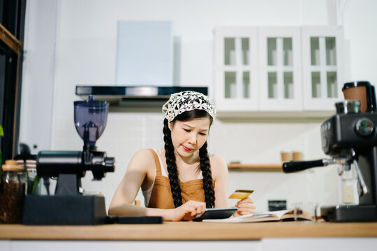 A worried female barista looks at a credit card while using a calculator at the counter. Concept of small business debt, financial stress
