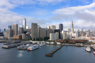 Obraz premium San Francisco's urban skyline stretching along the waterfront, featuring iconic towers, bustling piers, and boats docking at the Embarcadero under a partly cloudy morning sky
