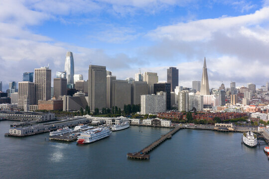 San Francisco's urban skyline stretching along the waterfront, featuring iconic towers, bustling piers, and boats docking at the Embarcadero under a partly cloudy morning sky