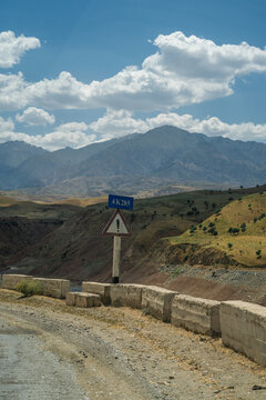 View of a road sign marked 4K285 and a warning sign along a concrete barrier with arid mountains and a blue sky in the background in Uzbekistan.