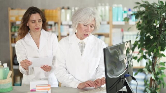Female pharmacists and computer at work counter, professional searches for data on vitamins to improve immunity, monitors warehouse stocks. In drug store worker type and using personal computer.