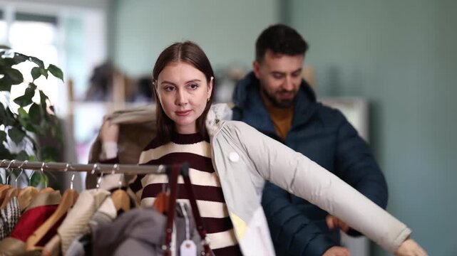Couple try on down jacket in front of mirror in clothes store. Family mutual time shopping. Buyer is immersed in choice of clothes. Determine comfort of clothes by fitting
