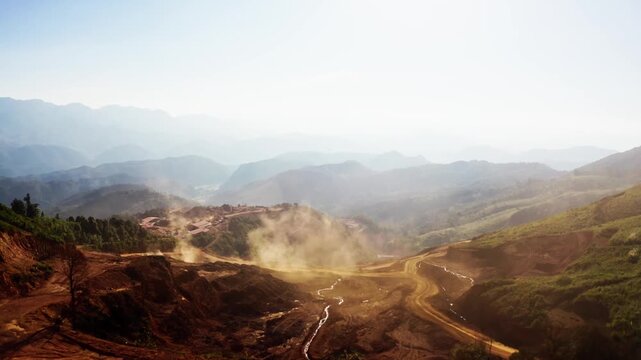 Top-down aerial tracking shot of an active iron ore mine, Red laterite soil and mining equipment contrast with surrounding green forest and mountains in Xieng Khouang province, Lao PDR.