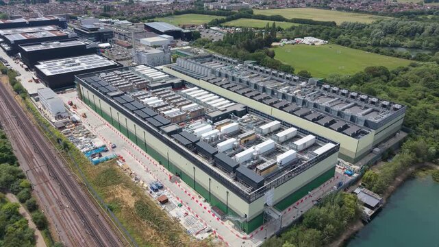 Aerial view of a large data center facility with industrial cooling units on the roof, situated near a railway line and a lake in London, London, United Kingdom.