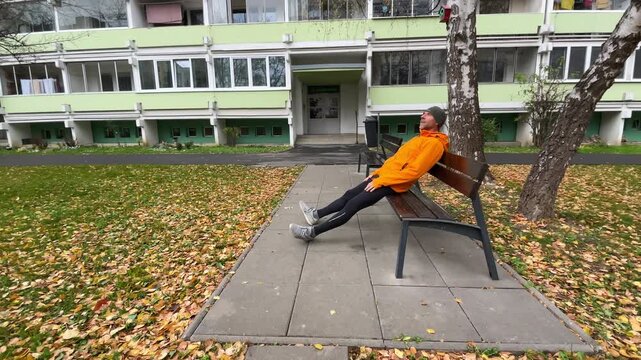 running man in orange waterproof jacket resting on the bench. Runner is dressed in athletic gear, showcasing active lifestyle in serene natural setting 4K video