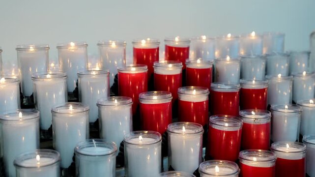 Rows of lit red and white prayer candles glowing inside a church or chapel. Peaceful spiritual atmosphere with memorial candles burning during religious worship.