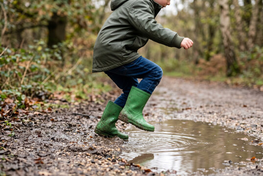 Boy in green rubber boots jumping over a woodland puddle on a muddy trail for outdoor activity and kids play background