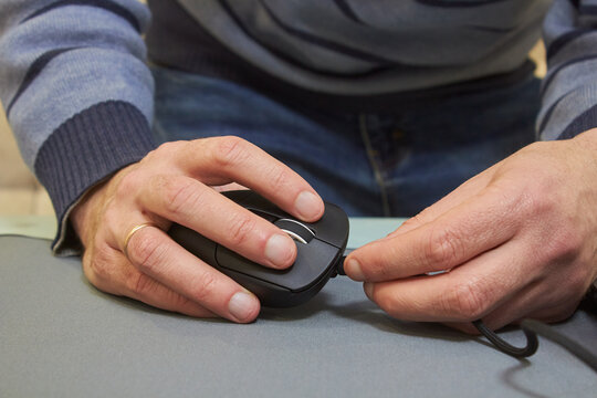 A man connects a cable to a computer mouse, Close-up of a man's hands in a gray sweater holding a black computer mouse and connecting the cable to it