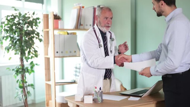 Elderly physician stands and shakes hand with adult man patient after consultation