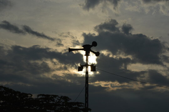 Silhouette of vintage outdoor air raid siren loudspeaker horn system on steel pole against dramatic cloudy sunset sky, public warning broadcast emergency alert system