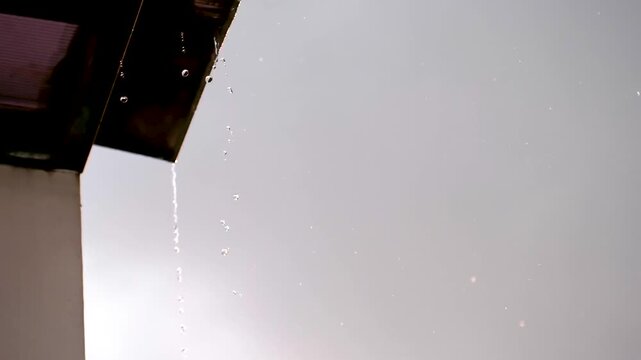 Rainwater dripping from a building roof during rainfall against a bright overcast sky. Weather-related scene showing water runoff and seasonal rain.