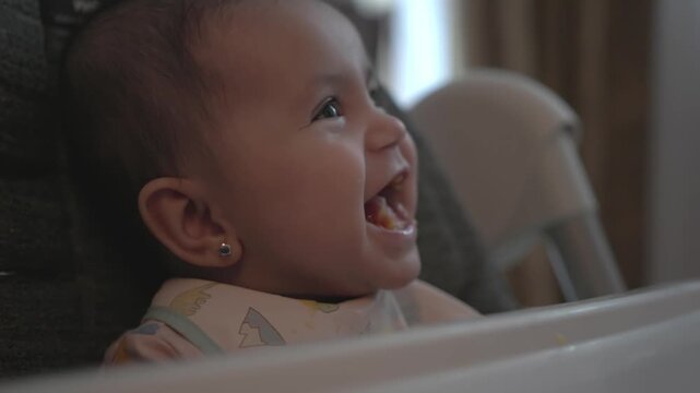 Close-up of a happy baby laughing in a high chair during mealtime at home, ideal for parenting, baby food, childcare and family lifestyle themes.