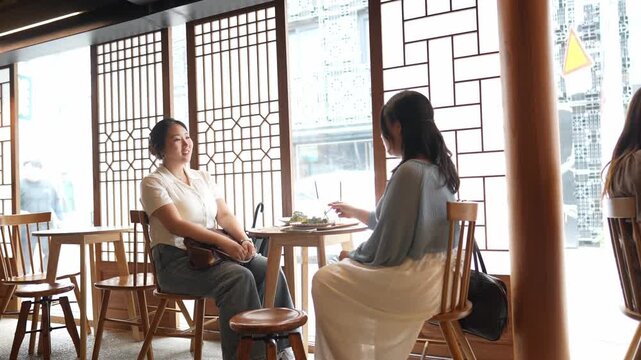 Two women sit at a wooden table. They chat, smiling, in a bright cafe. Large windows frame their conversation. Wooden chairs and lattice panels surround them. Soft light bathes the room, creating calm