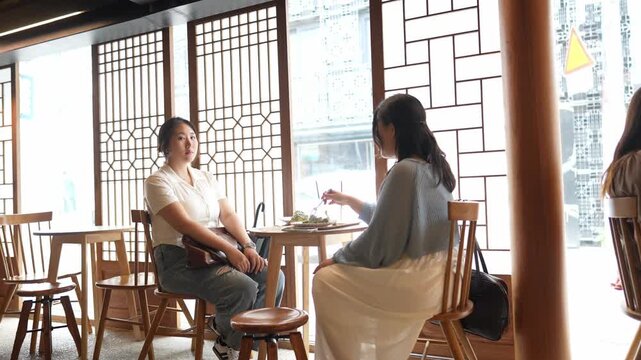 Two women chat near a window with wooden lattice. Soft light bathes a calm, quiet cafe setting. One smiles, the other listens attentively. Ideal for cozy cafe or friendship scenes