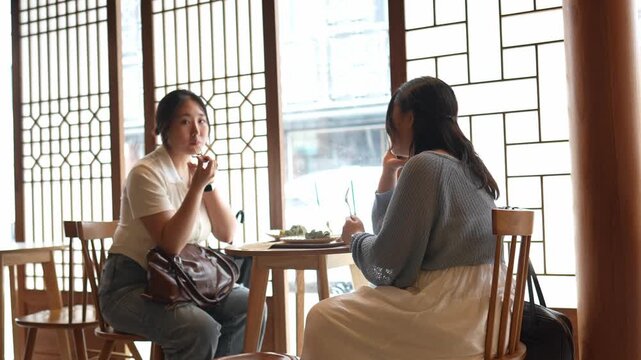 Two women share a quiet meal at a wooden table. They sit near large, patterned windows that let in soft light. One woman smiles, her eyes meet the other's gaze