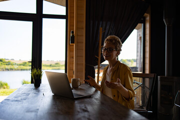 Naklejka premium A Caucasian woman with glasses smiles while gesturing during a video call on her laptop at a wooden table with a scenic lake view