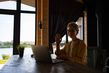 Naklejka premium A smiling Caucasian woman with glasses waves hello while on a video call on her laptop, seated at a wooden table with a scenic view outside