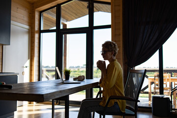Naklejka premium A Caucasian woman enjoys a hot beverage while working on her laptop in a modern wooden cabin with large windows overlooking a scenic landscape