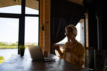 Naklejka premium A Caucasian woman wearing headphones and glasses smiles while working on her laptop at a wooden table with a scenic view outside the window