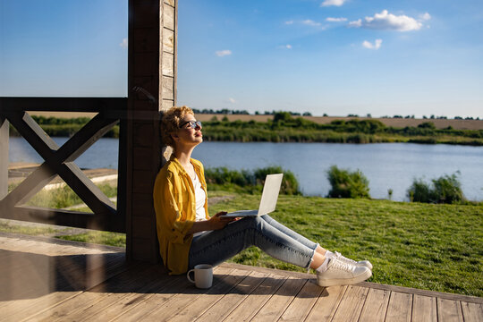 A Caucasian woman enjoys a sunny day while working on her laptop on a wooden porch overlooking a tranquil lake and green landscape