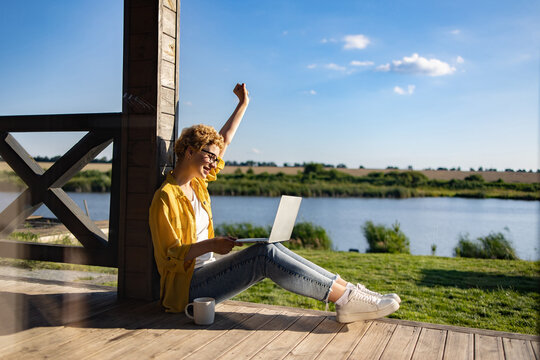A happy woman with short blonde hair and glasses raises her fist in triumph while working on her laptop on a wooden porch overlooking a serene lake and fields