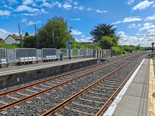 Fototapeta premium Train station with dual railway tracks extending into the distance. The tracks are bordered by gravel, and the platform has safety markings. Metal railings line the platform edge, with a few benches f