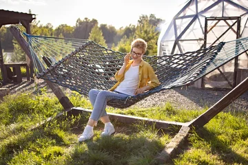 A young Caucasian woman with short blonde hair and glasses is talking on her phone while relaxing in a hammock on a sunny day in a grassy yard with a geodesic dome structure in the background © Sofiia Shunkina