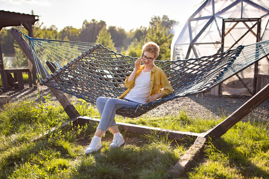A young Caucasian woman with short blonde hair and glasses is talking on her phone while relaxing in a hammock on a sunny day in a grassy yard with a geodesic dome structure in the background