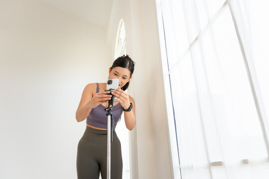 Young fitness woman setting up smartphone on tripod for recording workout, Influencer adjusting mobile phone on stand for fitness video content to stream yoga class at home in a bright studio