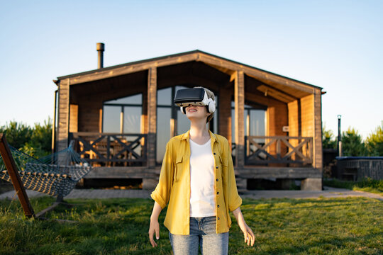 A young woman wearing a virtual reality headset and headphones enjoys an immersive experience in the yard of a wooden cabin
